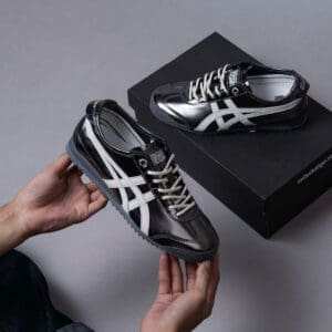 Black glossy sneakers with white stripes held in hands, a black shoebox in the background (Onitsuka Tiger style).]