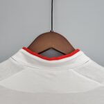 15f31af6_d624a319-0d21-4e9d-a1e2-74b9ee22647b | UAE SPORT STORE Close-up of a white shirt with red neckline on a wooden hanger against a gray background. Hanger supports a white shirt with red trim at the neck, seen from below.