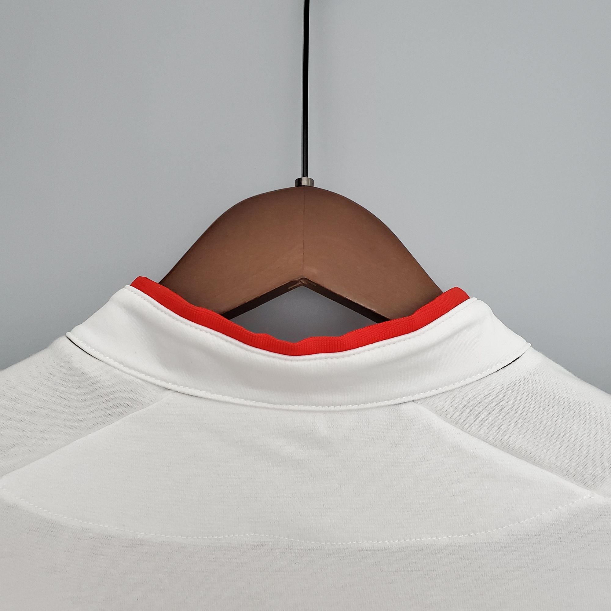 15f31af6_d624a319-0d21-4e9d-a1e2-74b9ee22647b | UAE SPORT STORE Close-up of a white shirt with red neckline on a wooden hanger against a gray background. Hanger supports a white shirt with red trim at the neck, seen from below.