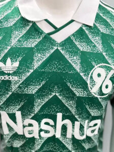 Close-up of a green geometric Adidas jersey with a white collar, Adidas logo on the left, sponsor text 'Nashua' across the chest, and a partial circular '96' emblem on the right.