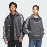 Two people wear matching gray Adidas jackets with white stripes, standing side by side; man in black shirt left, woman in jeans right.