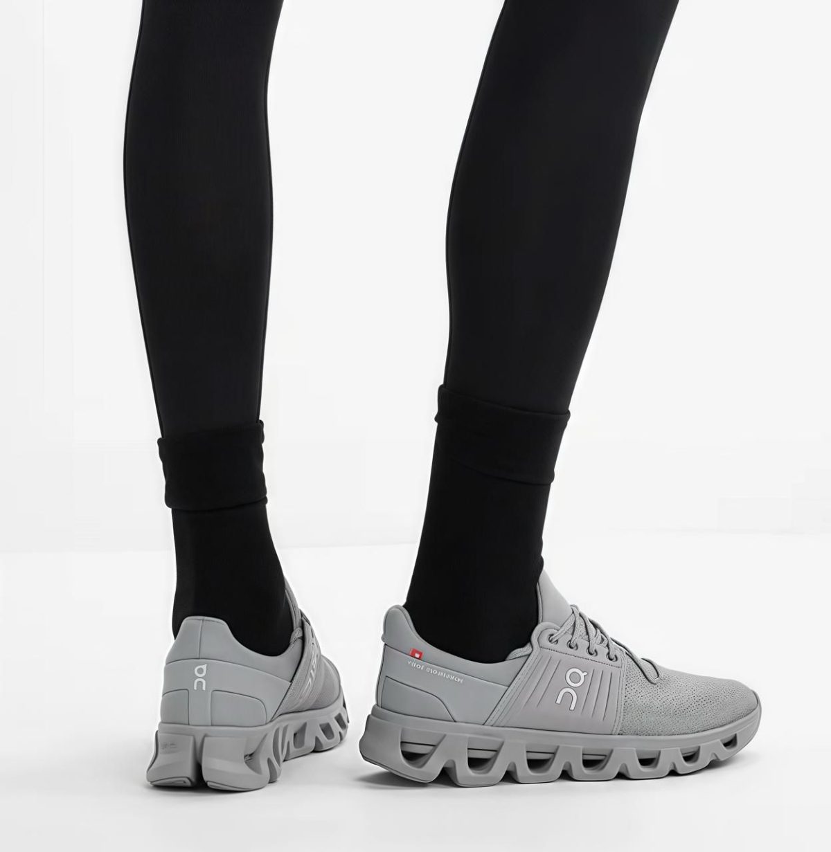 Close-up of legs in black leggings wearing light gray On running shoes with chunky, wavy soles on a white floor.