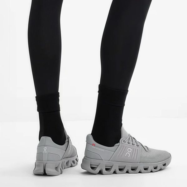Close-up of legs in black leggings wearing light gray On running shoes with chunky, wavy soles on a white floor.