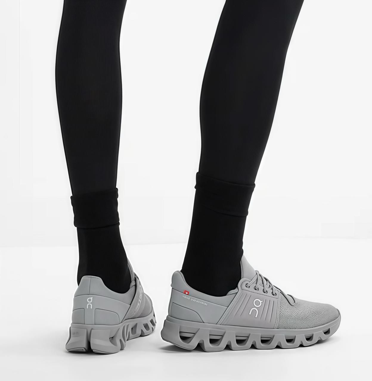 165e1cbe-23bb-4e7a-9726-8bc24c9faf2b | UAE SPORT STORE Close-up of legs in black leggings wearing light gray On running shoes with chunky, wavy soles on a white floor.