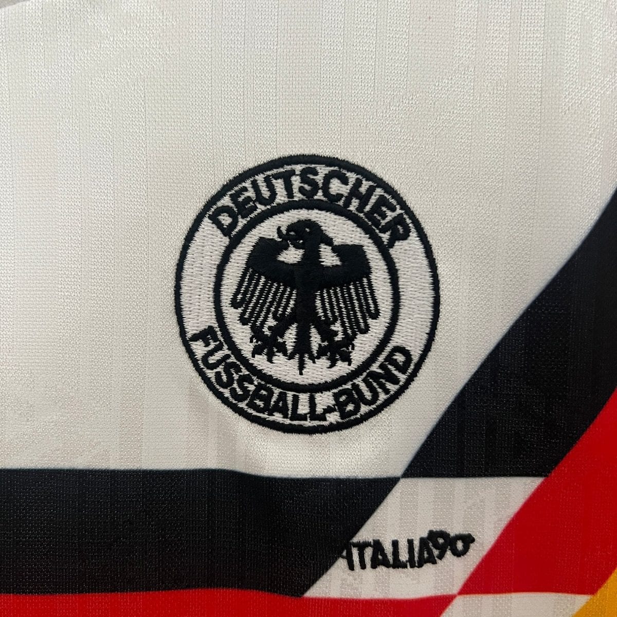 Close-up of a white fabric with a circular black-and-white embroidered patch showing an eagle and German text, over a diagonal black, white, and red stripe reminiscent of the Italian flag.