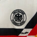 16602f93 | UAE SPORT STORE Close-up of a white fabric with a circular black-and-white embroidered patch showing an eagle and German text, over a diagonal black, white, and red stripe reminiscent of the Italian flag.