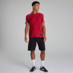 Male model wearing a red pinstripe soccer jersey with white piping, black shorts, white socks, and black sneakers against a neutral backdrop.