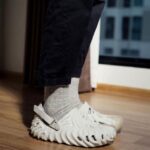 16a0a985-17ca-4ecf-a56d-de1a517bdf17 | UAE SPORT STORE Person wearing gray socks and white wave-patterned sandals with a back strap, standing on a wooden floor indoors near a window.