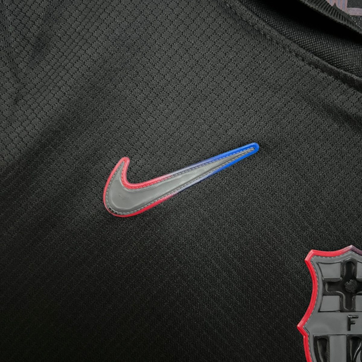Close-up of dark woven fabric with a partial view of a gray item featuring red and blue stitching at the bottom-right corner.
