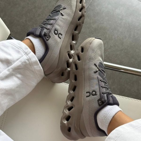 Pair of gray athletic sneakers with a segmented, hollow midsole, worn with white socks and light-colored pants while resting legs on a white surface.