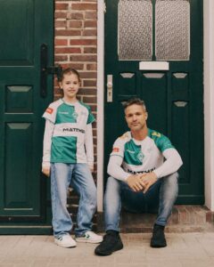 Girl and man in matching green-and-white soccer jerseys pose on a doorstep outside a brick building with green doors; girl stands while man sits on the step.