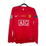 Red Manchester United long-sleeve jersey on a hanger showing the club crest, white Nike swoosh, and large AIG sponsor logo on the front; UEFA patch on the left sleeve and small text/details visible at the bottom hem.