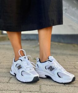 17b6f77a-d4ec-4d31-b4e6-662bd10ed202 | UAE SPORT STORE Close-up of white New Balance athletic sneakers worn by a person in a black skirt, standing on a paved surface.