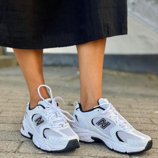 Close-up of white New Balance athletic sneakers worn by a person in a black skirt, standing on a paved surface.