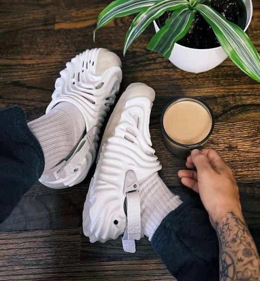 17ff5213-4f05-41e9-bfe4-5adfdaa5da80 | UAE SPORT STORE Person wearing white sneakers and light socks rests feet on a dark wood floor, holding a mug of coffee; a potted plant sits nearby.