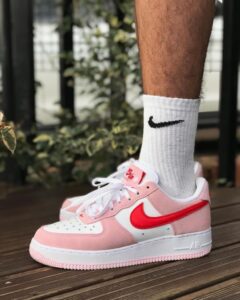 Close-up of a pink and white Nike Air Force 1 sneaker on a wooden deck, worn with a white sock featuring a black Nike swoosh.