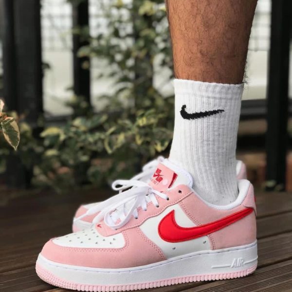 Close-up of a pink and white Nike Air Force 1 sneaker on a wooden deck, worn with a white sock featuring a black Nike swoosh.