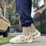 Person wearing beige socks and cream perforated sandals stepping on a sidewalk, with a parked car in the background.