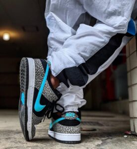 Person wearing black, gray, and turquoise Nike sneakers with elephant print, standing with one foot raised on a concrete sidewalk.