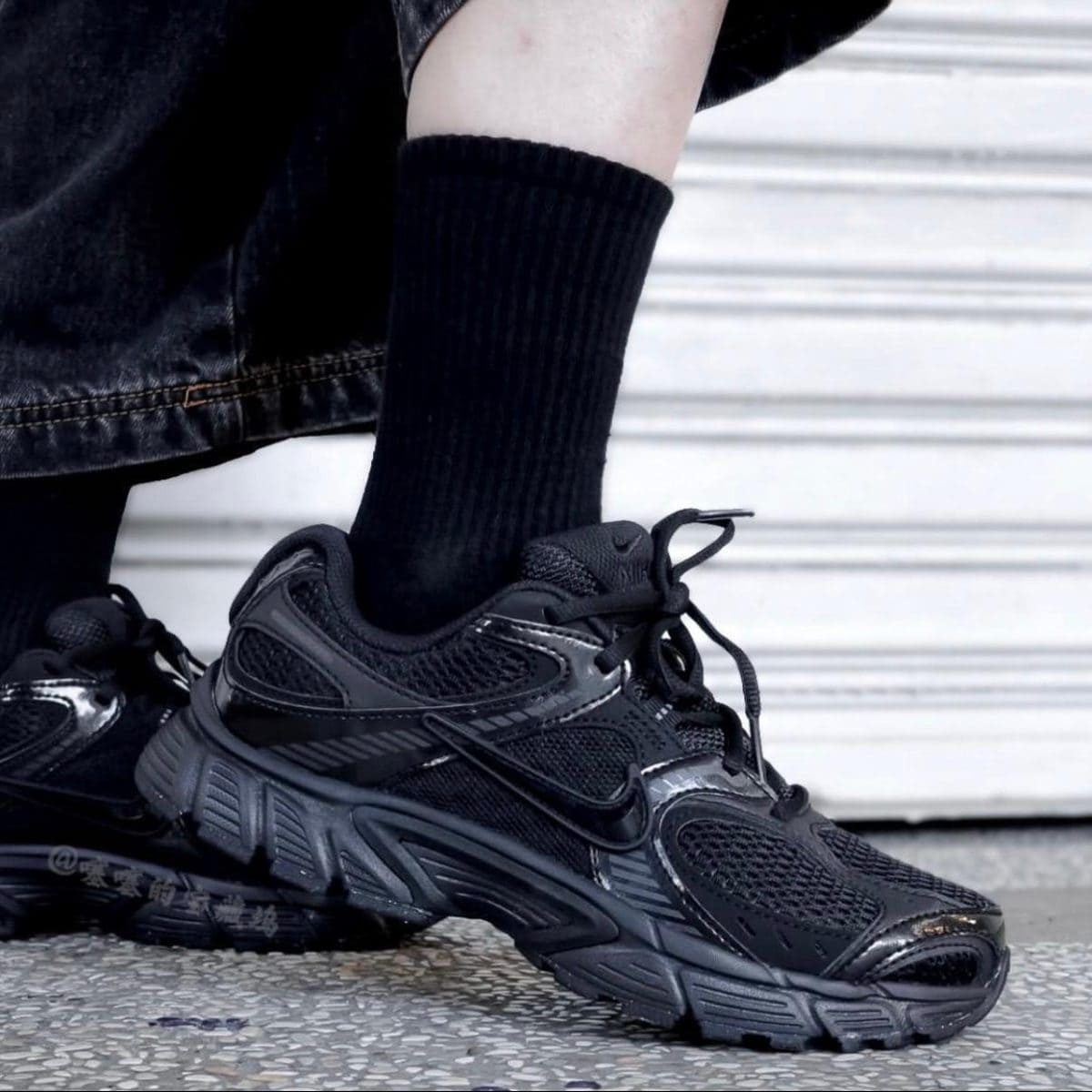 Close-up of a black chunky sneakers on a person wearing a black sock, standing on a textured pavement with a light backdrop.