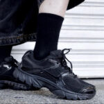 Close-up of a black chunky sneakers on a person wearing a black sock, standing on a textured pavement with a light backdrop.