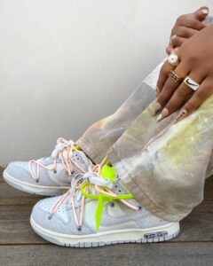 Feet in gray sneakers with pink and neon yellow laces rest on a wooden floor; hand with gold rings rests beside them.