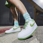 Person wearing white Nike sneakers with lime green swoosh and sole, mint socks, with leg tattoos, stepping on a concrete sidewalk outdoors.