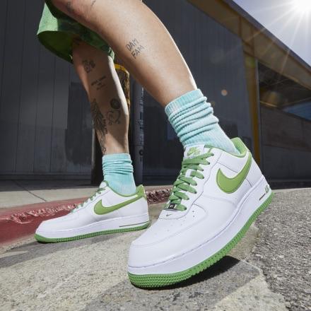 Person wearing white Nike sneakers with lime green swoosh and sole, mint socks, with leg tattoos, stepping on a concrete sidewalk outdoors.