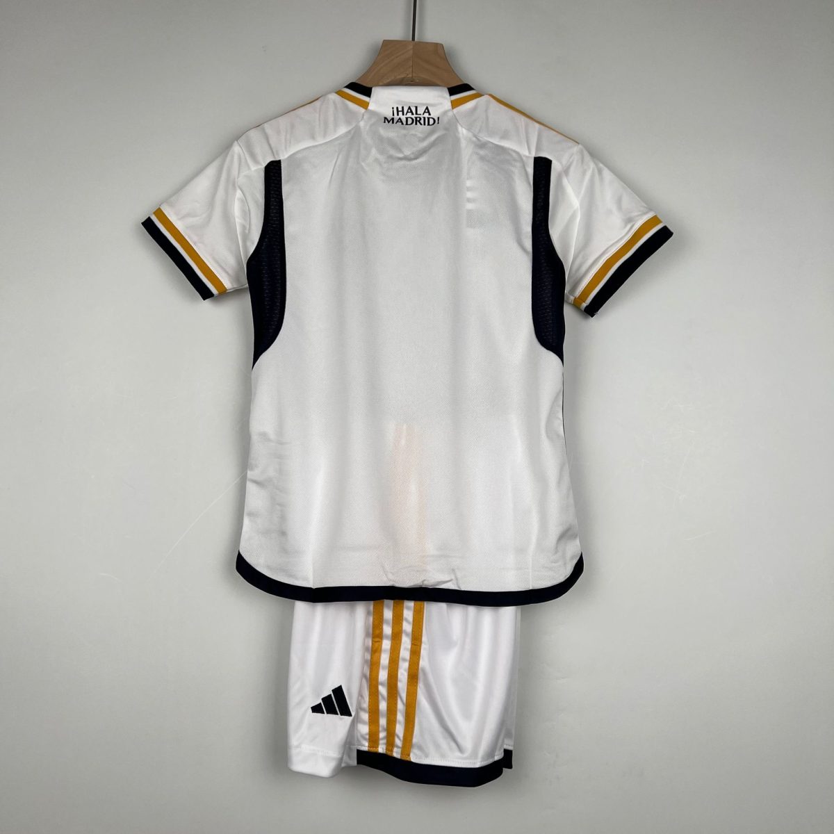 Back view of a white Adidas soccer jersey hanging on a wooden hanger, with black and gold trim and the text ¡HALA MADRID! on the upper back.