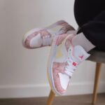 Pastel pink and white high-top sneakers with a yellow swoosh, resting on a chair leg against a light wall.