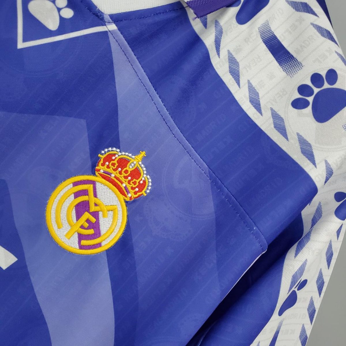 Real Madrid crest stitched on a blue sports jersey with white geometric patterns in the background, near the chest area.