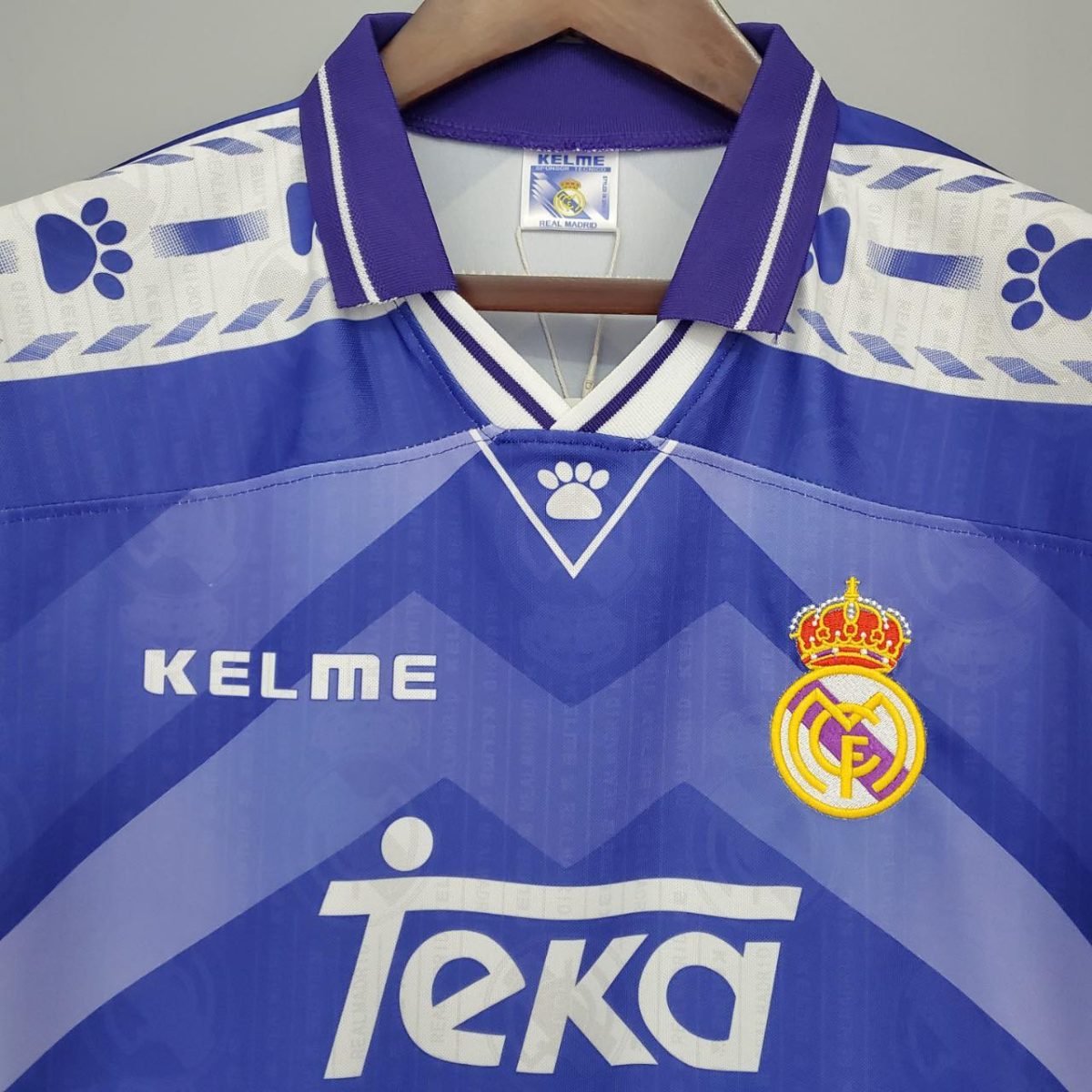Blue Real Madrid football jersey with purple trim on a hanger; features the KELME brand and Real Madrid crest on the chest area, with a large sponsor logo across the front.