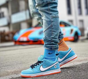 Close-up of blue Nike sneakers with white swoosh, orange outsole, and rolled jeans on a city street with a colorful car in the background.