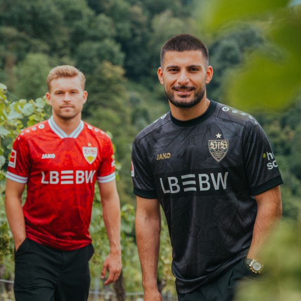 Two men in red and black soccer jerseys stand in a vineyard, posing and smiling toward the camera.