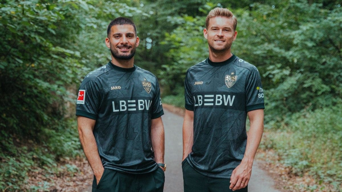 Two men in dark soccer jerseys stand side by side on a forest path, smiling at the camera.
