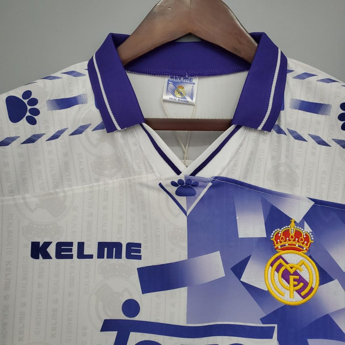 White Real Madrid football jersey with blue geometric accents and crest on the chest, hanging on a wooden hanger on a gray background