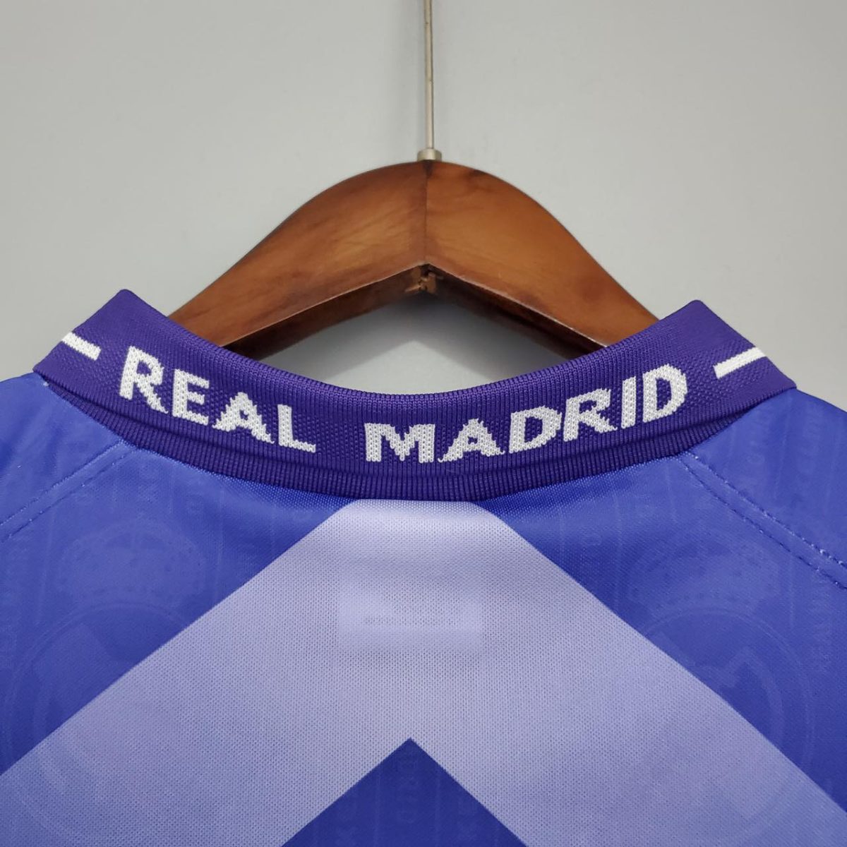 Blue Real Madrid football jersey hanging on a wooden hanger, close-up of the collar with white 'REAL MADRID' text.