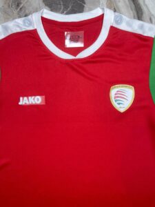 Red athletic jersey with a white collar, JAKO logo on the left chest, and a crest badge on the right chest featuring red/green swooshes and Arabic text; green side panel visible on the right edge.