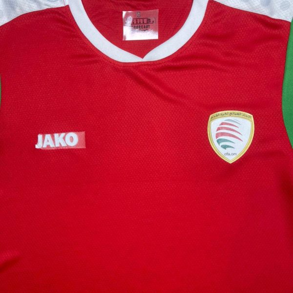 Red athletic jersey with a white collar, JAKO logo on the left chest, and a crest badge on the right chest featuring red/green swooshes and Arabic text; green side panel visible on the right edge.