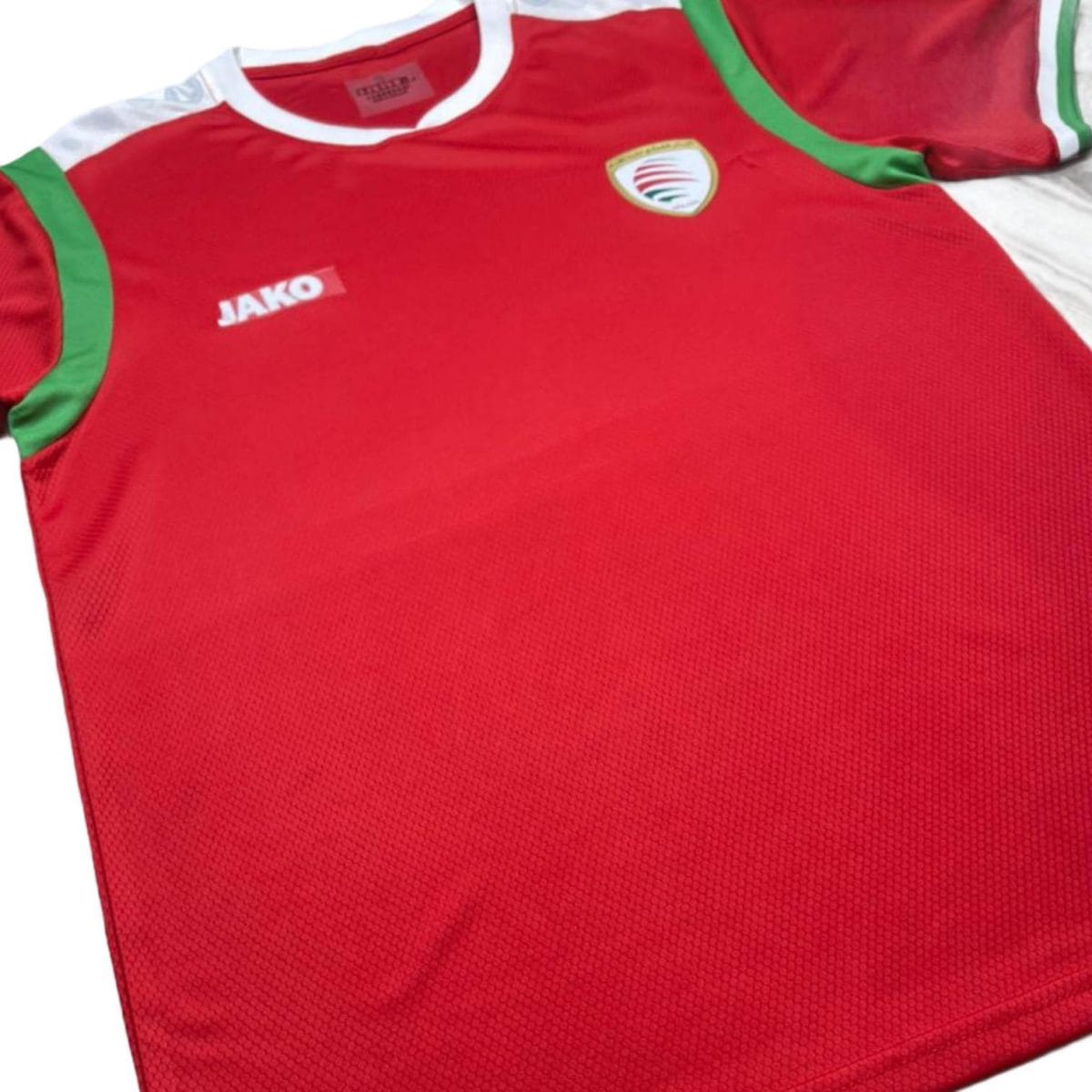 Red athletic jersey with white collar and green piping; features a JAKO logo on the chest and a small circular crest badge.