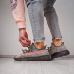 Person tying the laces of gray patterned knit sneakers with thick brown soles, wearing light gray frayed-hem jeans on a light floor.