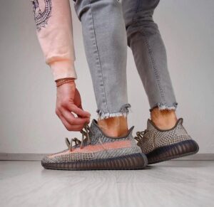 Person tying the laces of gray patterned knit sneakers with thick brown soles, wearing light gray frayed-hem jeans on a light floor.