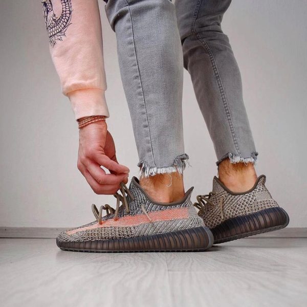 Person tying the laces of gray patterned knit sneakers with thick brown soles, wearing light gray frayed-hem jeans on a light floor.