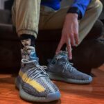 Person seated, tying light blue knit sneakers with a yellow stripe while wearing cartoon-pattern socks and khaki pants on a wooden floor.
