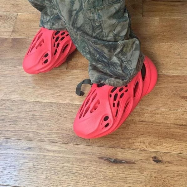 Person wearing bright red Crocs with multiple ventilation holes, standing on a wooden floor in camouflage pants.