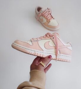 Pink and white Nike sneakers; one shoe is held in a hand while a matching pair rests in the background against a light backdrop.