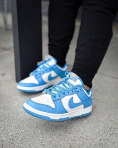 Pair of blue and white Nike sneakers on a gray sidewalk, laces tied, wearing black pants nearby.