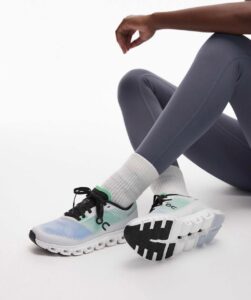 Close-up of white and teal running shoes with chunky sole, worn with gray leggings and white socks, on a white floor.