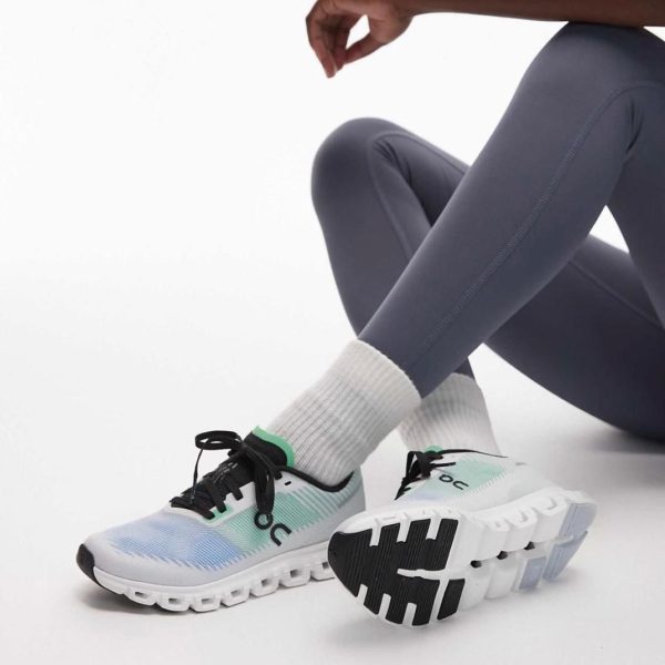 Close-up of white and teal running shoes with chunky sole, worn with gray leggings and white socks, on a white floor.