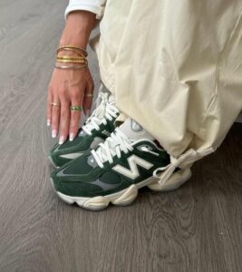 Person wearing green and white New Balance sneakers, beige rolled-up pants, resting hand with gold bracelets on the shoe.